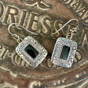 May include: A pair of silver-toned drop earrings. Each earring features a rectangular black center stone surrounded by small, clear crystals and decorative silver filigree. The earrings are displayed on a textured, brown surface.