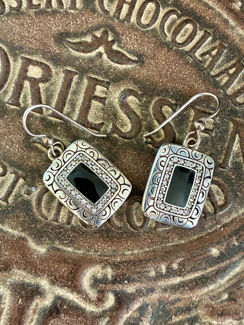 May include: A pair of silver-toned drop earrings. Each earring features a rectangular black center stone surrounded by small, clear crystals and decorative silver filigree. The earrings are displayed on a textured, brown surface.