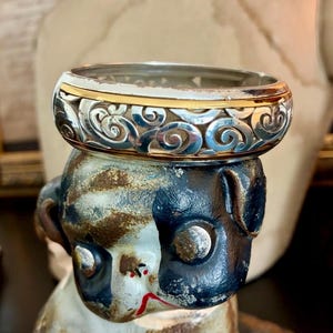 Puede incluir: Un anillo plateado con un intrincado dise&ntilde;o de remolinos y una banda dorada. El anillo se exhibe sobre una figura decorativa con una combinaci&oacute;n de colores negro, blanco y marr&oacute;n. El dise&ntilde;o del anillo es detallado y llamativo.