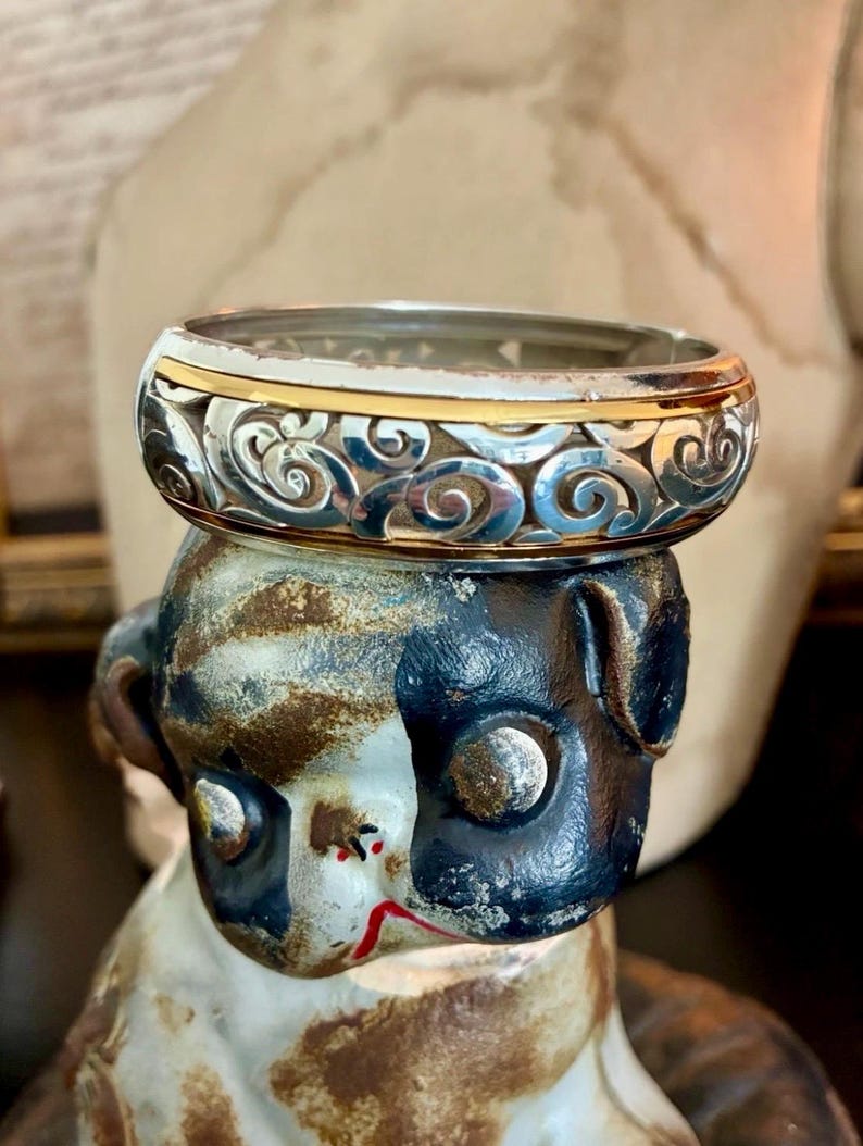Puede incluir: Un anillo plateado con un intrincado dise&ntilde;o de remolinos y una banda dorada. El anillo se exhibe sobre una figura decorativa con una combinaci&oacute;n de colores negro, blanco y marr&oacute;n. El dise&ntilde;o del anillo es detallado y llamativo.