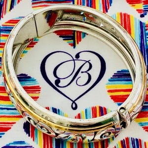 Puede incluir: Pulsera articulada plateada y dorada con adornos de volutas y un logotipo en forma de coraz&oacute;n. La pulsera se encuentra sobre un fondo colorido de corazones y rayas, creando un contraste vibrante.