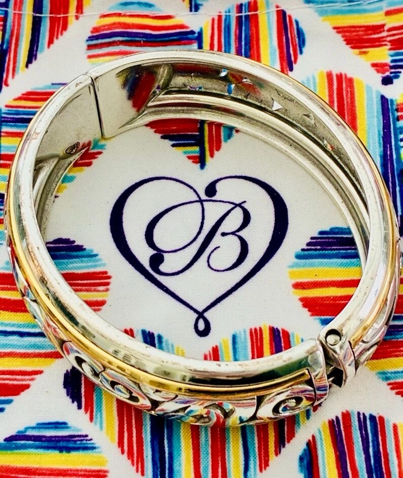 Puede incluir: Pulsera articulada plateada y dorada con adornos de volutas y un logotipo en forma de coraz&oacute;n. La pulsera se encuentra sobre un fondo colorido de corazones y rayas, creando un contraste vibrante.