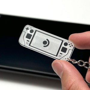 May include: A clear plastic keychain shaped like a handheld video game console with a black and white design. The keychain has a silver metal ring.