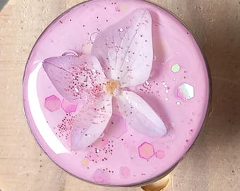 May include: A pink resin bottle stopper with a dried flower and glitter details, set on a round wooden base. The stopper is a decorative item for bottles. The wooden base is approximately 5 cm in diameter.