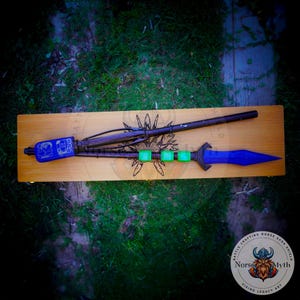 May include: A blue and black spear with a wooden handle and green accents. The spear is displayed on a wooden board with the text "Battle Crafting Horse Saga Shield Norse Myth Viking Legacy Art".