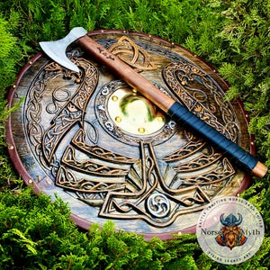 May include: A detailed wooden Viking shield with carved patterns and a brass centre, accompanied by a wooden-handled axe. The shield has a brown and gold design with a leather edge. The axe features a silver blade and a black grip. The image includes the text "Norse Myth".