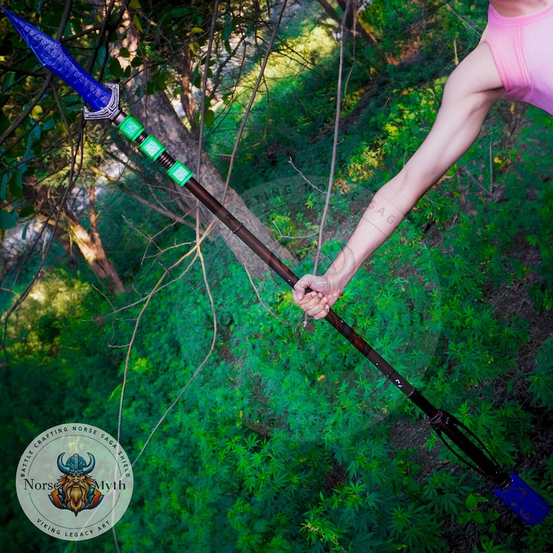 May include: A blue and green spear with a wooden shaft and a leather strap. The spear is held by a person's hand in a forest setting. The spear has a green and silver design on the shaft and a blue blade with a silver tip. The spear is labeled "Battle Crafting Norse Saga Shield Norse Myth Viking Legacy Art".