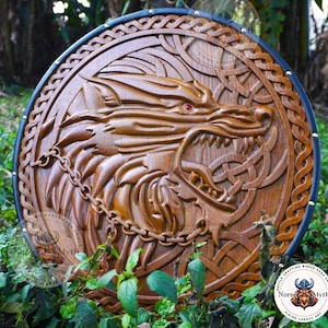 May include: A round, wooden shield with a carved wolf design. The shield is brown with a black border and features a detailed carving of a wolf's head with red eyes and a chain. The shield has a Viking theme.