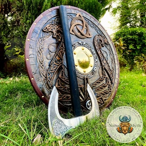 Könnte beinhalten: Ein Holzschild mit aufwendigen Schnitzereien eines Drachen und einem Knotenmuster. Der Schild ist mit Messingakzenten verziert und hat einen Lederriemen. Eine silberne Axt mit einem geschnitzten Stiel lehnt am Schild. Schild und Axt befinden sich auf einer grünen Grasfläche. Der Text "Norse Myth" ist auf einem runden Aufkleber sichtbar.
