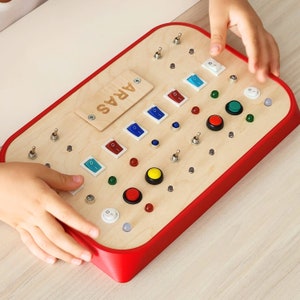 Può includere: Un pannello di gioco in legno con bordo rosso e vari interruttori, pulsanti e luci. Il pannello è etichettato "ARAS".