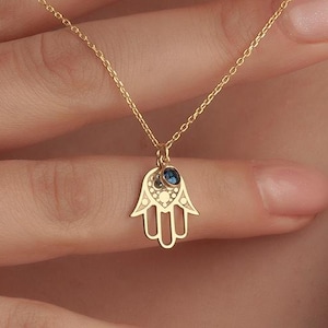 Könnte beinhalten: Eine goldfarbene Halskette mit einem Hamsa-Hand-Anhänger. Der Anhänger hat einen blauen Edelstein und kleine klare Steine. Die Halskette ist zierlich und besteht aus einer dünnen Kette. Der Hintergrund ist ein weißes Hemd.