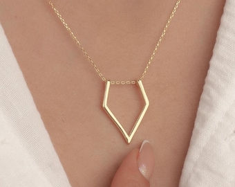 May include: A gold-colored necklace with a geometric pendant in a diamond shape. The pendant hangs from a delicate chain. The necklace is displayed on a person wearing a white shirt.