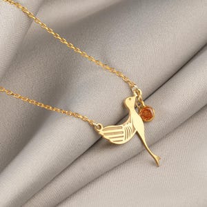 May include: A gold-coloured necklace with a bird pendant and a small, round, orange gemstone. The bird has outstretched wings and a long tail. The necklace chain is delicate and gold-toned.