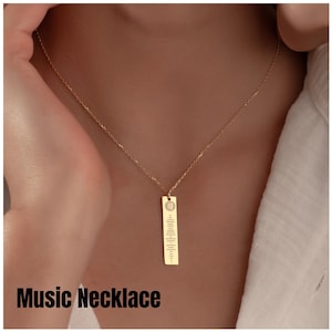 Könnte beinhalten: Eine goldfarbene Musik-Halskette mit einem rechteckigen Anhänger. Der Anhänger zeigt oben ein Musiksymbol und eine Reihe von Linien, die einen Musikcode darstellen. Die Halskette wird auf der Brust einer Person getragen. Der Text "Music Necklace" steht unten.