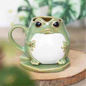 May include: A ceramic frog-shaped mug with a green and white glaze. The mug has a handle and sits on a green lily pad-shaped base. The frog's eyes are black, and the mug is on a wooden surface.