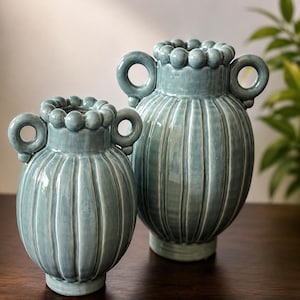 May include: Two light blue ceramic vases with a glossy finish, featuring vertical ribbed textures, circular side handles, and a decorative beaded rim around the top opening. The vases are rounded in shape with a narrow base and a wider body.