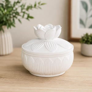 May include: A matte white ceramic lidded jar featuring a sculpted lotus flower handle on the lid. The exterior of both the lid and the rounded base is decorated with a repeating embossed petal pattern, creating a textured, uniform appearance.