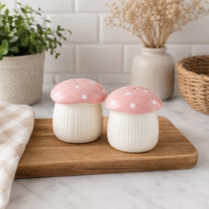 May include: A pair of ceramic salt and pepper shakers shaped like mushrooms. Each shaker features a rounded, light pink cap with white polka dots and a cream-coloured, vertically ribbed base. The shakers are positioned side by side on a wooden board.