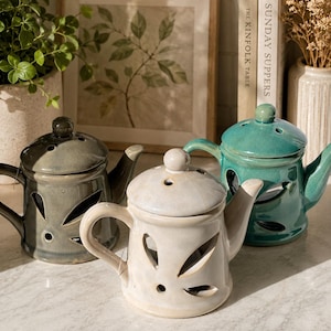 May include: Three ceramic teapots with lids, each featuring a glossy finish and leaf-shaped cutouts. One teapot is light grey, one is cream-coloured, and one is teal. Each teapot has a curved handle, a spout, and a small knob on the lid.