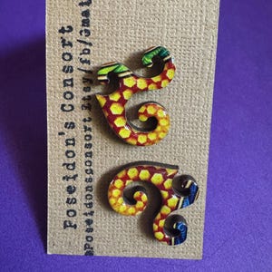 May include: Two decorative, swirled earrings with a red and yellow honeycomb pattern, displayed on a tan card. The earrings have a glossy finish and are accented with green and blue details. The card has the text "Poseidon's Consort" and social media information.