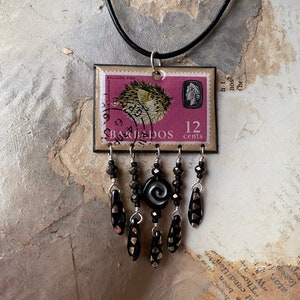 May include: A black cord necklace with a pendant featuring a vintage Barbados postage stamp with a picture of a balloon fish and the text "BALLOON FISH" and "BARBADOS 12 cents". The pendant has black beads and a silver spiral charm hanging from it.