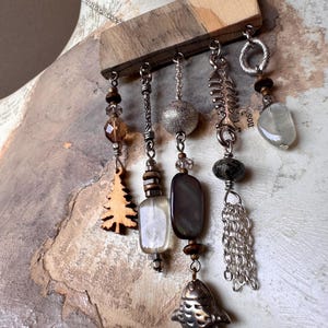 May include: A necklace featuring a rectangular wooden bar suspended from a silver chain. Five dangling charms hang from the bar, including a wooden tree, a silver fish skeleton, a metal fish, various glass beads, and a silver chain tassel.