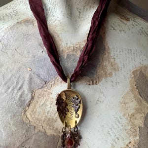May include: A unique necklace with a burgundy ribbon. The pendant is a gold-toned spoon shape with leaf accents and the word "Camilla" engraved. A cluster of decorative elements and a red crystal dangle from the bottom.