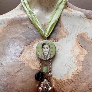 May include: A necklace featuring a light green fabric ribbon and a circular pendant with a black and white illustration of a person surrounded by the number 5. Below the pendant hang silver chains, a green bead, a black circular charm, and a gas mask.