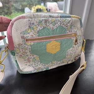 May include: A rectangular crossbody bag crafted from quilted fabric featuring a patchwork of floral patterns and a central hexagonal flower motif in mint green and yellow. The bag includes a gold-tone zipper, matching hardware, and a beige strap.