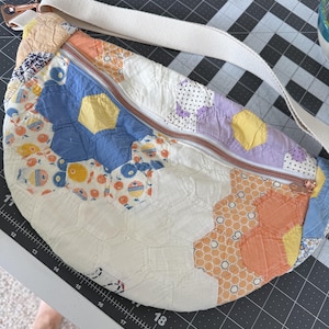 May include: A crescent-shaped fanny pack constructed from a patchwork of quilted fabric hexagons in shades of blue, yellow, orange, and white. The bag features a top zipper closure and an adjustable off-white woven strap with metal hardware.