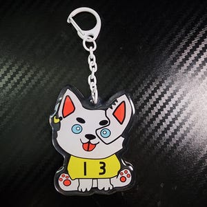 Puede incluir: Un llavero de acrílico transparente con un perro de dibujos animados con cuerpo blanco, orejas rojas y ojos azules. El perro lleva una camisa amarilla con el número "13". El llavero tiene una cadena y un cierre blancos.