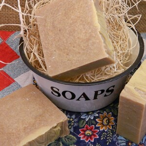 May include: Three bars of handmade soap in varying shades of brown and tan. One bar rests in a white enamel bowl labeled "SOAPS" and filled with straw. The soaps sit on a patchwork quilt.