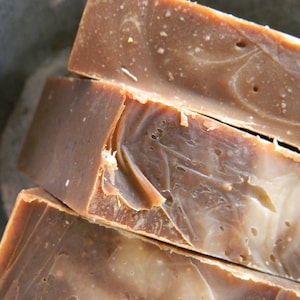 May include: Close-up of three bars of handmade soap. The soap has a brown color with lighter swirls and speckles throughout. The edges of the soap are slightly rough, indicating a natural, handcrafted product. The soap is likely for personal care.