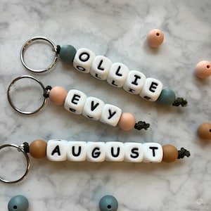 May include: Three personalised keychains with the names Ollie, Evy, and August spelled out in white letter beads. The beads are strung on a cord with alternating brown and blue beads. The keychains are on a white marble surface.