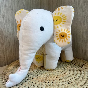 Könnte beinhalten: Ein weißes Stofftier-Elefant mit gelben Stoffohren, die ein Muster aus lächelnden Sonnen zeigen. Der Elefant sitzt auf einer geflochtenen Strohmatte.