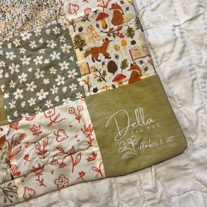 Peut inclure: Une courtepointe patchwork avec des carrés de tissu à motifs floraux et forestiers. La courtepointe présente un renard, des champignons et des motifs floraux dans des tons de vert, d'orange et de crème. Le coin porte la broderie "Della Rae Vos September 5, 2023".