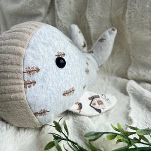 May include: A grey and white whale plush toy with brown tree designs. The whale has a brown knitted head and a black button eye.