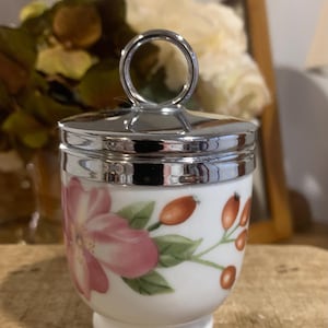 May include: A white porcelain egg coddler featuring a floral pattern with a large pink flower and orange berries on green stems. The container has a polished silver-tone metal lid with a circular ring handle at the top and a pedestal base.