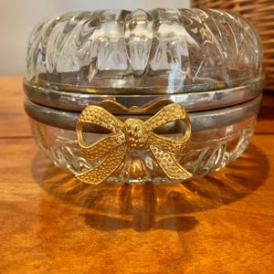May include: A clear glass trinket box with a silver rim and a gold bow clasp. The box has a ribbed design on the lid and base. The gold bow is in the center of the box.