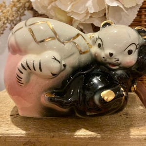 May include: A ceramic figurine of a bear in a reclining pose. The bear is primarily black and white with pink accents and gold detailing. The figurine has a vintage, cartoonish aesthetic.
