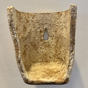 May include: A wall-mounted ceramic planter with a rough, speckled cream and brown textured finish. The vessel features a U-shaped opening and a single teardrop-shaped hole near the top center for mounting, showcasing a porous, stone-like appearance.