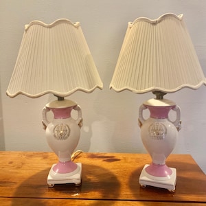 May include: Pair of decorative table lamps. Each lamp features a white ceramic base with pink accents and gold detailing. The lampshades are cream-colored and pleated. The lamps are placed on a wooden surface.