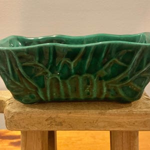 May include: A green ceramic planter with a leafy design. The planter is rectangular and has a wavy rim.