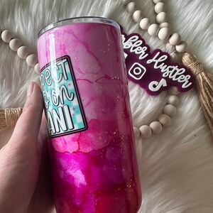 May include: A pink and magenta tumbler with a watercolor effect and gold flecks. The tumbler features a sticker that reads "Expect the Mani" in a retro font. The tumbler is held by a hand.