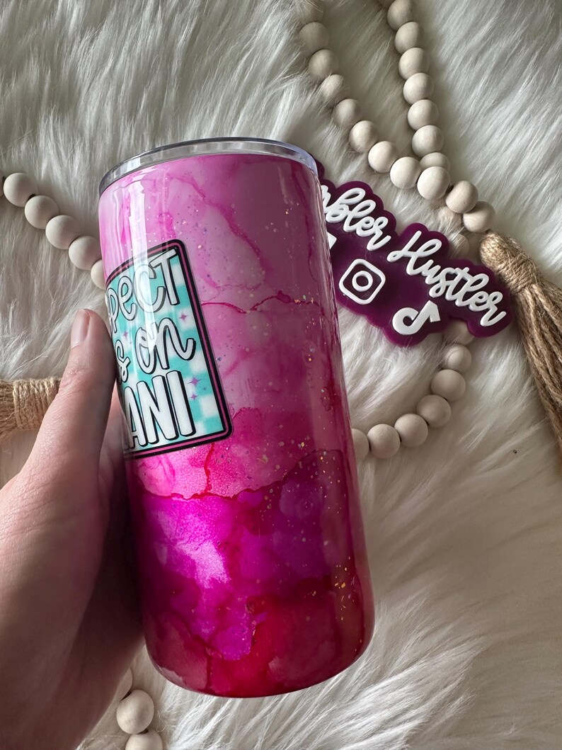 May include: A pink and magenta tumbler with a watercolor effect and gold flecks. The tumbler features a sticker that reads "Expect the Mani" in a retro font. The tumbler is held by a hand.
