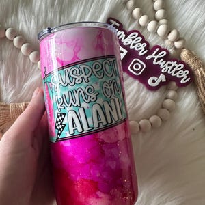 May include: A pink and magenta tumbler with a lid, featuring the text "Suspect Runs on Alani" in a turquoise and white design. The tumbler has a glittery finish and is held by a hand.