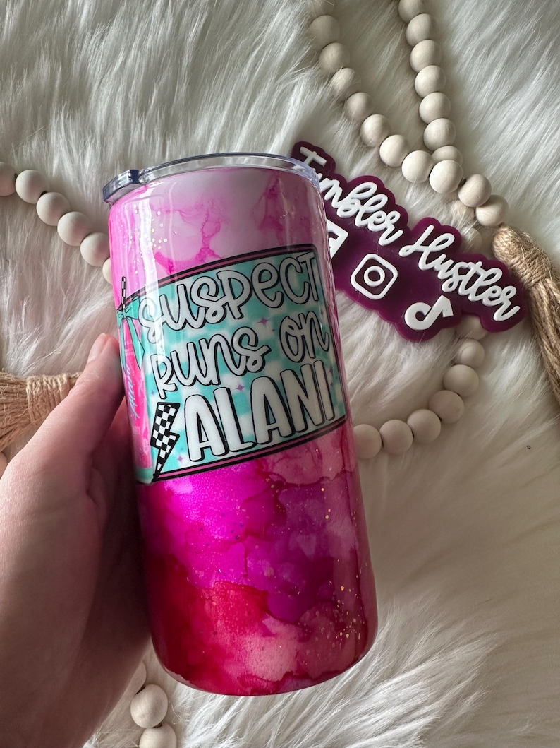 May include: A pink and magenta tumbler with a lid, featuring the text "Suspect Runs on Alani" in a turquoise and white design. The tumbler has a glittery finish and is held by a hand.