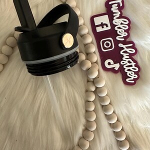 May include: Black water bottle lid with a flip-top spout and a handle. The lid has a straw and a gold-colored button. A maroon sticker with white text that says "Tumbler Hustler" and social media icons is also visible.