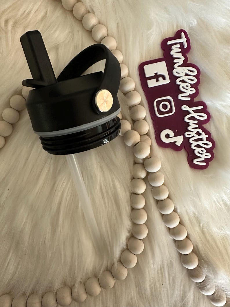 May include: Black water bottle lid with a flip-top spout and a handle. The lid has a straw and a gold-colored button. A maroon sticker with white text that says "Tumbler Hustler" and social media icons is also visible.