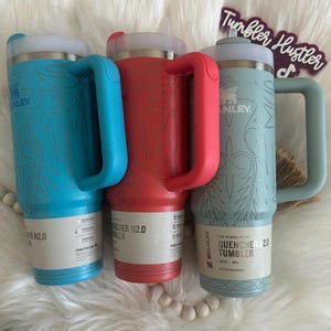 Może przedstawiać: Trzy kubki Stanley Quencher H2.0 w kolorze turkusowym, czerwonym i morskim. Każdy kubek ma pasujący uchwyt i pokrywkę z dekoracyjnym kwiatowym wzorem. Kubki mają pojemność 887 ml.